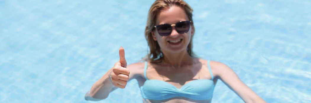 Portrait of happy young woman swim in pool and show thumbs up from water. Relaxing atmosphere on vacation, sunny day. Holiday, resort, hotel, fun concept