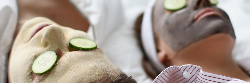 Close-up of woman and man friends resting on bed with facial mask and cucumbers. Cute couple make treatment for skin on weekend. Wellness, skincare concept