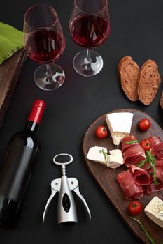 Above overhead view flat lay still life of assortment various cheese and delicatessen with traditional bread and a bottle of red wine with two glasses on a old wooden board on black table. Top view.