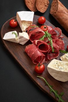 Cheese, prosciutto, bread, vegetables and spices on wooden board on black background with copy space. Still life. Top view. Flat lay