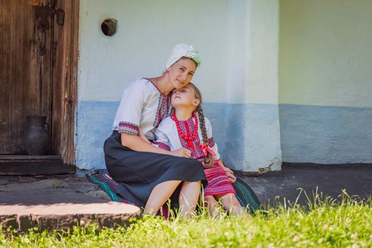 mother and daughter in Ukrainian national costumes are sitting near an old house