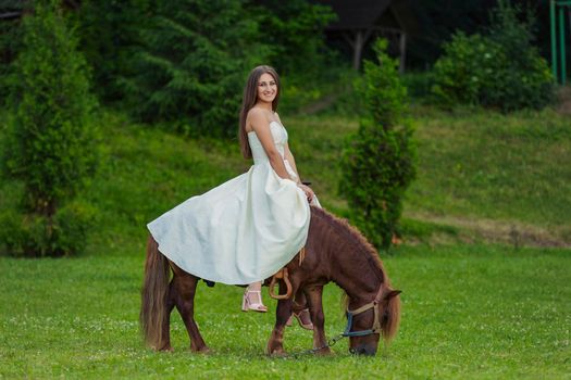 girl in a white dress rides a pony on a green lawn
