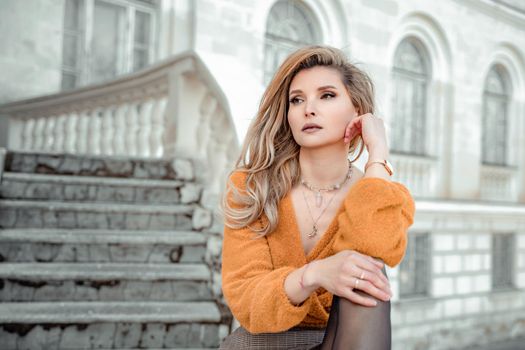 A middle-aged woman looks like a good blonde with curly beautiful hair and makeup on the background of the building. She is wearing a yellow sweater