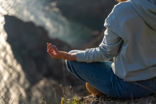 Woman tourist enjoying the sunset over the sea mountain landscape. Sits outdoors on a rock above the sea. She is wearing jeans and a blue hoodie. Healthy lifestyle, harmony and meditation.