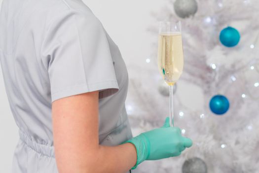 Beautiful young female doctor in protective gloves with glass of champagne standing near the christmas tree. Soft focus