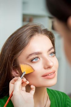 Professional make up artist applying powder by brush on cheeks of beautiful young caucasian woman in beauty salon