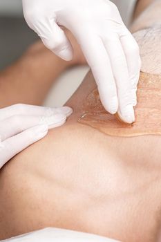 Epilation chest of young male with liquid wax paste by hands of female cosmetologist in beauty salon