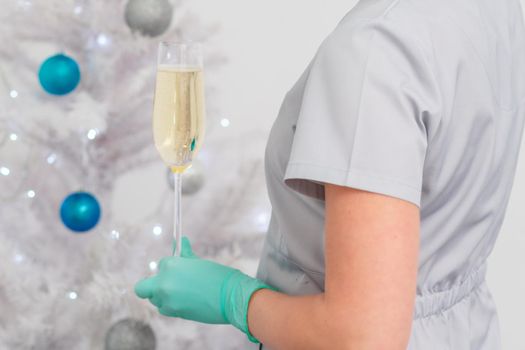 Beautiful young female doctor in protective gloves with glass of champagne standing near the christmas tree