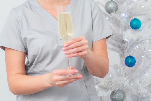 Beautiful young female doctor with glass of champagne standing near the christmas tree. Blurred
