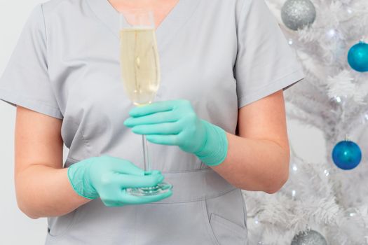 Beautiful young female doctor in protective gloves with glass of champagne standing near the christmas tree