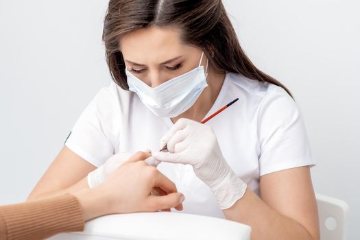 Manicure master with protective mask applying transparent nail polish on female nails in beauty salon