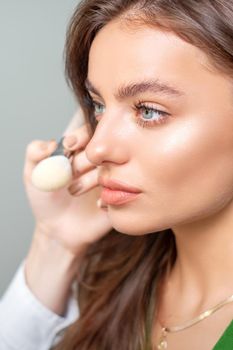 Makeup artist applying dry cosmetic tonal foundation on the face of young woman using brush tool in beauty salon