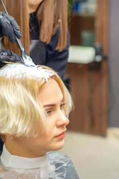 Hairdresser in black gloves dyes hair of young woman in white color in hairdress salon