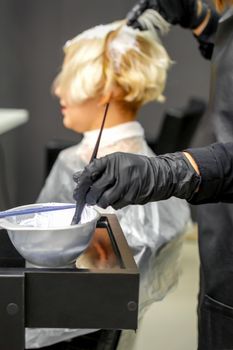 Hairdresser in black gloves dyes hair of young woman in white color in hairdress salon. Soft focus