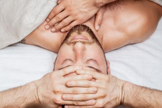 Handsome young man receiving relaxing head massage by four hand of two masseurs in health spa center