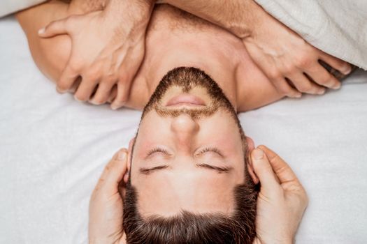 Handsome young man receiving relaxing head massage by four hand of two masseurs in health spa center