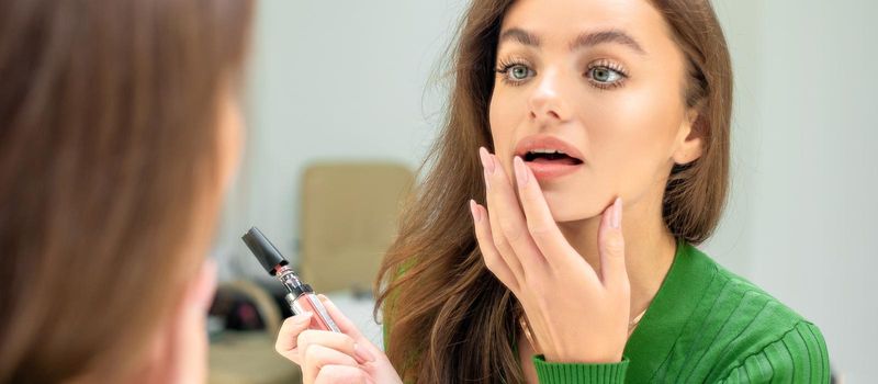 Beautiful young caucasian woman applies lipstick on her lips with her finger looking in mirror