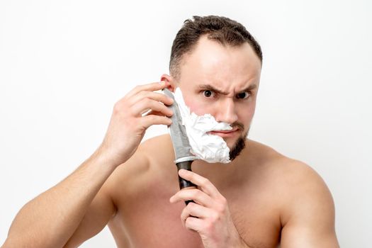 Young caucasian man shaving beard with knife on white background