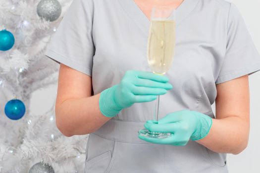 Beautiful young female doctor in protective gloves with glass of champagne standing near the christmas tree. Defocused