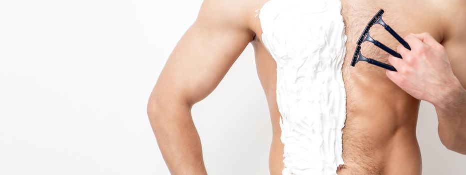 Young caucasian man with beard holds razor shaves his chest with white shaving foam on white background. Man shaving his torso