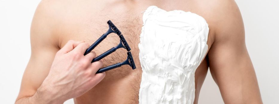 Young caucasian man with beard holds razor shaves his chest with white shaving foam on white background. Man shaving his torso