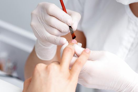 Manicure master in rubber gloves applying transparent nail polish on female nails in beauty salon