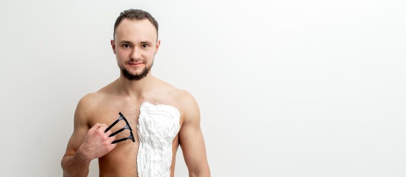 Young caucasian man with beard holds razor shaves his chest with white shaving foam on white background. Man shaving his torso