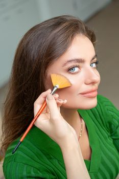 Professional make up artist applying powder by brush on cheeks of beautiful young caucasian woman in beauty salon