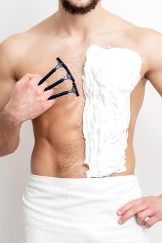 Young caucasian man with beard holds razor shaves his chest with white shaving foam on white background. Man shaving his torso