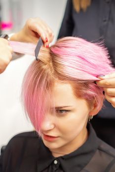 Hairdresser straights pink hair of young woman by hair iron in hairdress salon