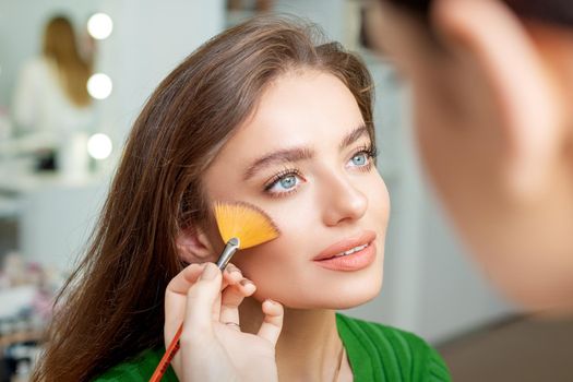 Professional make up artist applying powder by brush on cheeks of beautiful young caucasian woman in beauty salon