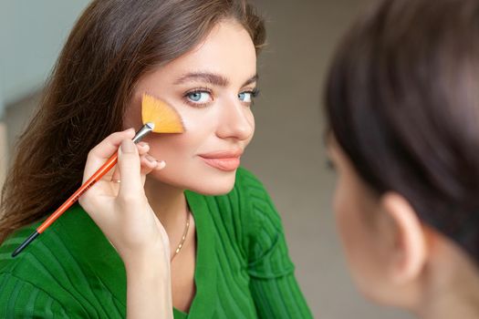 Professional make up artist applying powder by brush on cheeks of beautiful young caucasian woman in beauty salon