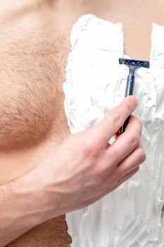 Young caucasian man with beard holds razor shaves his chest with white shaving foam on white background. Man shaving his torso