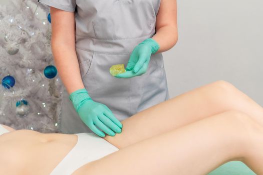 Cosmetologist in gloves applying paste for sugaring depilation on legs of young woman. Christmas discount waxing