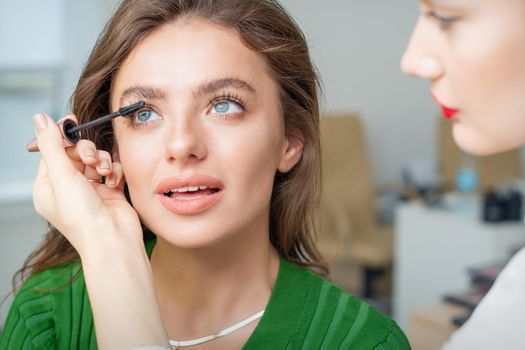 Makeup artist applies mascara on eyelash of young caucasian woman in beauty salon