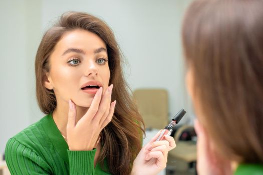 Beautiful young caucasian woman applies lipstick on her lips with her finger looking in mirror