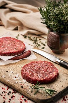 On a wooden cutting board on kraft paper there are raw beef burgers for burgers, spices, rosemary, a pepper mill, a cooking process. Top view. Copy space.