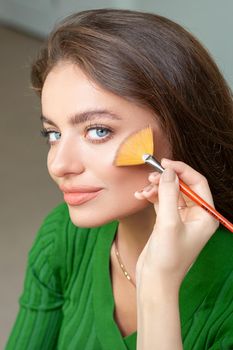 Professional make up artist applying powder by brush on cheeks of beautiful young caucasian woman in beauty salon