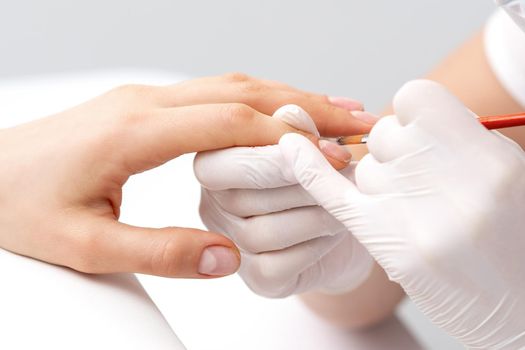 Manicure master in rubber gloves applying transparent nail polish on female nails in beauty salon