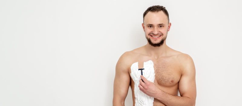 Young caucasian man with beard holds razor shaves his chest with white shaving foam on white background. Man shaving his torso