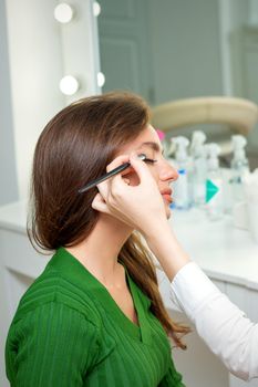 Makeup artist applies eyeshadow powder on eyes of young caucasian woman by brush tool in beauty salon