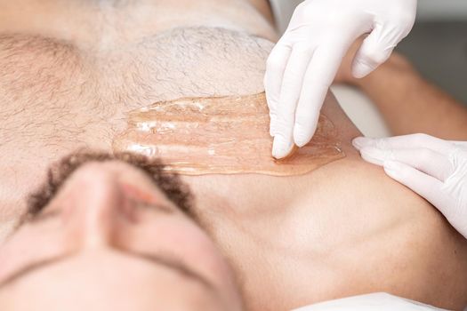 Epilation chest of young male with liquid wax paste by hands of female cosmetologist in beauty salon