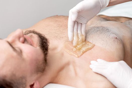 Epilation chest of young male with liquid wax paste by hands of female cosmetologist in beauty salon