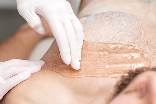 Epilation chest of young male with liquid wax paste by hands of female cosmetologist in beauty salon