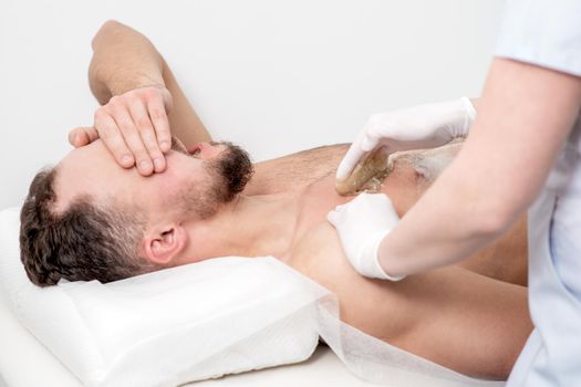 Epilation chest of young male with liquid wax paste by hands of female cosmetologist in beauty salon