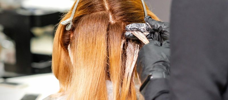 Back view of professional hairdresser dyes hair of young woman in beauty salon