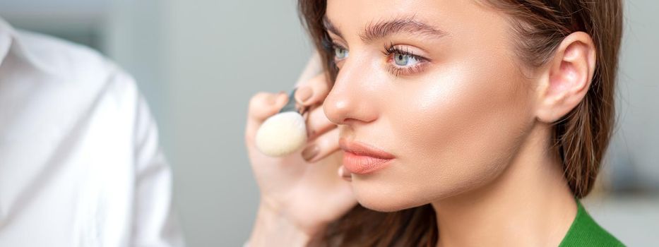 Makeup artist applying dry cosmetic tonal foundation on the face of young woman using brush tool in beauty salon