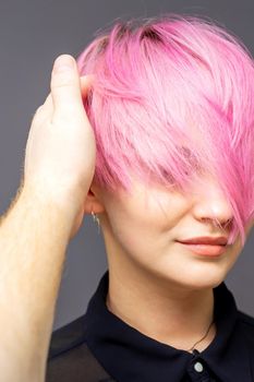 Hairdresser checking short pink hairstyle of young woman on gray background