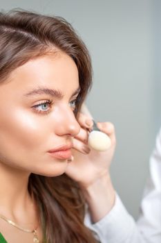 Makeup artist applying dry cosmetic tonal foundation on the face of young woman using brush tool in beauty salon