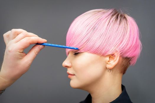 Hairdresser checking short pink hairstyle of young woman with comb on gray background